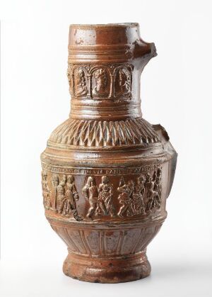  A stoneware jug titled "Bondedans" with a varied warm brown color palette. It has a flared rim, a curved handle, a relief of dancing figures around the body, and a geometric herringbone pattern on the base. Artist unknown.