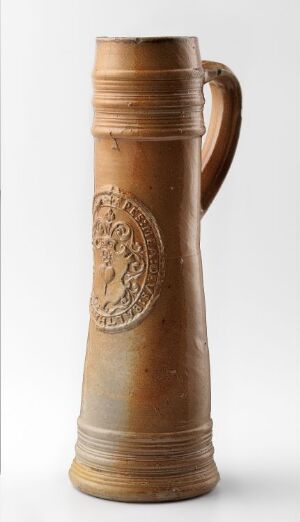  A tall, honey-brown ceramic beer stein with a glossy finish, featuring a lighter relief medallion on the front and a sturdy handle, set against a soft gray-to-white gradient background.