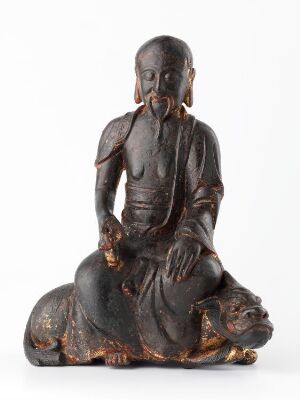  An intricately carved dark patina sculpture depicting an elderly figure with elongated earlobes sitting peacefully atop a mythical beast, possibly a lion, adorned with a draped robe and exuding an aura of wisdom and calm.