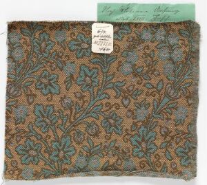  Historic brown textile fragment with an ornate pattern of blue, teal, and tan floral designs, featuring a small label with handwriting and partially covered by a green piece of paper in the upper right corner.