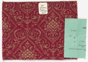  A rectangular swatch of intricate red patterned fabric with a white handwritten tag attached to the top left corner, and a separate teal-colored paper note with printed text and handwritten marks to the right, implying archival or historical contexts.