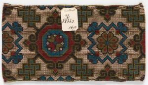  A small rectangular handwoven textile or rug with a traditional geometric and floral pattern. The design includes a central red diamond-shaped medallion with blue and white accents, flanked by two similar motifs, and an intricate border in shades of blue, green, and beige. An old paper tag with handwritten numbers is attached to one edge.