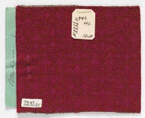  A photograph of a maroon fabric swatch with subtle woven patterns, presented on a backing card with a slim green strip on the left edge, and featuring two labels with handwritten and printed text for identification.