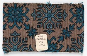  A rectangular, woven fabric with intricate geometric patterns in shades of blue on a muted brown background, featuring star and diamond motifs arranged in horizontal bands, and a small white label with handwritten text attached to the lower corner.