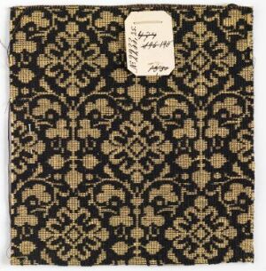  A square fabric swatch with a luxurious black, golden yellow, and beige ornate floral and geometric pattern, featuring a small white label with handwritten text attached to the top right corner.