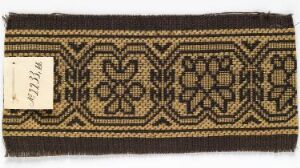  A section of woven fabric featuring a symmetrical pattern with black and gold colors, showcasing geometric and stylized floral designs, along with a small white inventory tag with numbers.