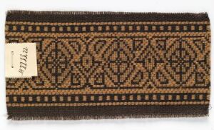  A rectangular textile with fringed edges and a brown and beige geometric pattern, featuring diamond shapes and chevrons, with a paper tag on one corner.