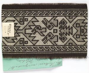  A woven textile fragment with a black base and intricate white geometric patterns, including a central diamond motif, alongside a white label and a strip of light blue paper with cursive writing.