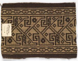  A woven textile with a symmetrical geometric pattern in black and beige tones, featuring diamond shapes and angular forms, indicative of traditional craftsmanship. Artist name and title unknown.