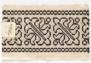  A textile fragment with intricate black embroidery on an off-white background, featuring a symmetrical floral or foliate design, bordered by a geometric frame, and a small, unreadable label attached to the lower left corner.