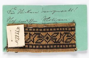  A small piece of woven beadwork with a geometric pattern predominantly in gold, brown, and white, resting on a textured green background with cursive handwriting and a small cream-colored tag with numbers. Artistname and title are unknown.