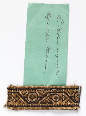  A light teal piece of paper with diagonal cursive handwriting is placed above a smaller piece of textile with a black, gold, and white geometric pattern and fringed edges, both on a white background. Artist name and title are unknown.