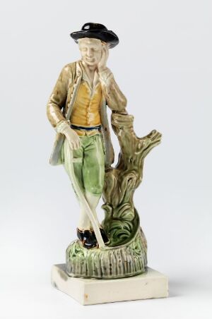  Ceramic statue of a man in traditional mustard-yellow shirt and green trousers, with his right hand tipping a black wide-brimmed hat, standing next to a textured tree stump on a square off-white base.