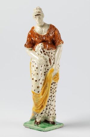  A figurine of a woman with a serene expression, wearing a burnt orange blouse, dotted white skirt, holding a mustard yellow shawl, standing on a green base.