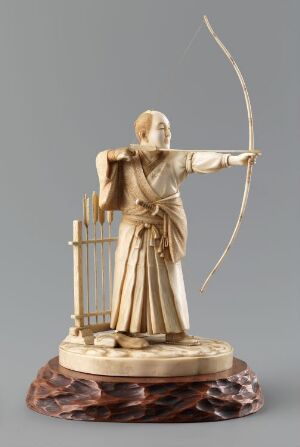  An ivory-colored sculpture of an archer in traditional attire, with a detailed depiction of flowing garments and a drawn bow, set upon a dark, patterned base against a neutral gray background.