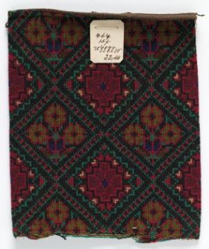  A fragment of a woven textile with an intricate geometric pattern in deep reds, dark greens, golds, and hints of blue, featuring an aged tag with handwriting in the upper right corner.