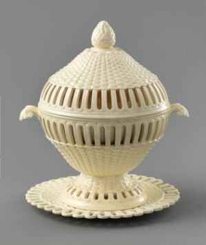  An intricately designed ivory-colored porcelain spherical object with a domed lid, featuring a pattern of radiating lines and small circular openings, complete with ear-shaped handles and resting on a matching scalloped base plate. Artist name and title are unknown.