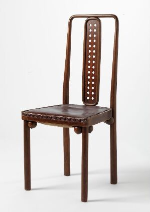  An elegant chair designed by Josef Hoffmann made from turned and steam-bent beech wood, featuring a vertically elongated backrest with neatly arranged circular perforations and a seat covered with dark brown oilcloth, set against a plain light grey background.