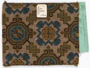  A photograph of a small textile fabric sample with a detailed geometric pattern in dark teal, deep brown, and lighter blue on a light beige background, accompanied by an attached white tag and partial view of a note card with handwritten text.