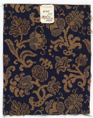  A piece of fabric with a rich navy blue background and golden brown floral and foliate pattern, with a white label in the upper left corner displaying text and numbers, suggesting an item from a curated or historical collection. Artist name and title unknown.