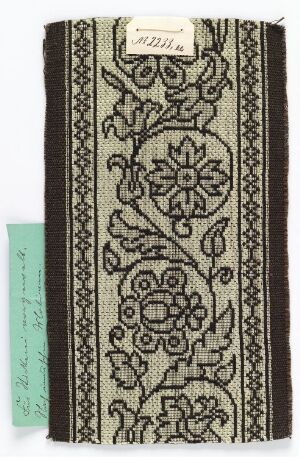  A rectangular fabric with dark brown and beige symmetrical floral patterns, framed by vertical borders, and bearing a small white label in the corner and a green cardboard tag on the left side.