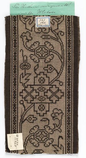  An antique brown textile fragment with dark brown floral and geometric patterns, bordered by thin dark stripes, and a pale mint green band at one end with a paper label attached.