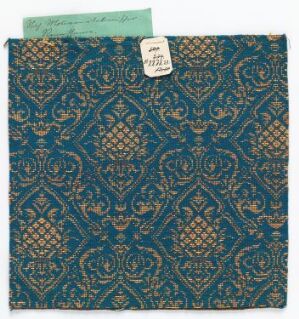 
 A swatch of vintage textile featuring an elaborate teal and metallic gold pattern with a partial light green paper tag in the top left corner, indicating cataloging details.