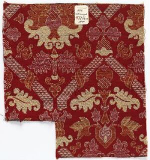  A piece of deep red fabric with ornate golden-tan patterns in a diagonal grid layout, showing a mixture of stylized floral and geometric shapes, with a white label in the top right and a cut corner revealing the fabric's structure.