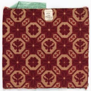  A square piece of deep red fabric adorned with symmetrical gold geometric and floral patterns, featuring a contrasting teal swatch with an attached label in the upper left corner.