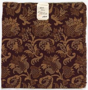  A square swatch of dark brown fabric adorned with a lighter brown paisley and botanical print. A white tag is affixed to the top left corner with a handwritten number "777;B" followed by "ab."