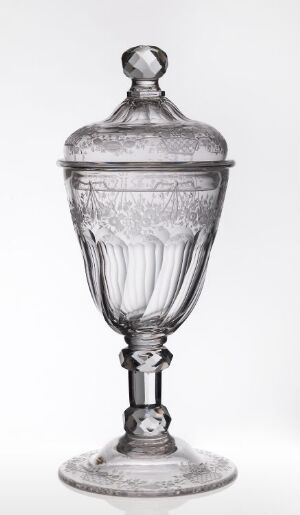  An elegantly designed, transparent glass jar with a lidded top and faceted knob, featuring textured patterns on its curved body, a decorative stem, and a stable base, set against a light background.