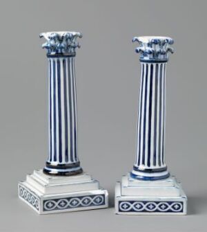  Two classical column-style candlesticks in white and cobalt blue, set against a soft grey background. The candlesticks feature fluted shafts, decorative acanthus leaf capitals, and square, stepped bases with a blue looped motif.