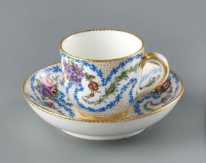 
 An ornate porcelain teacup and saucer with a white base color, decorated with vibrant floral patterns in shades of purple, pink, blue, and green, and adorned with rich cobalt blue swirls and gold trimmings. The cup is positioned in the saucer at an angle that showcases the interior and exterior designs.