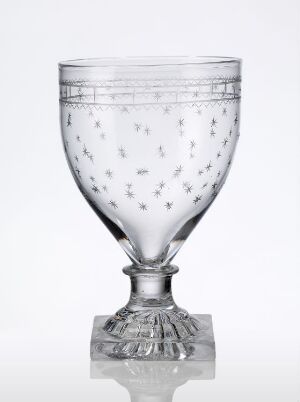  An elegant, clear glass goblet with a faceted stem and air bubbles embedded in the bowl, placed on a reflective surface against a light gray background. The glass is clear, and the goblet has a broad bowl and a solid base, presenting a minimalist and refined aesthetic. Artist name and title are unknown.