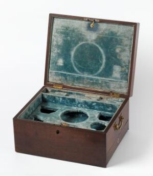  An antique wooden box with brass hardware, partially opened to reveal a faded blue-lined interior with various circular compartments.