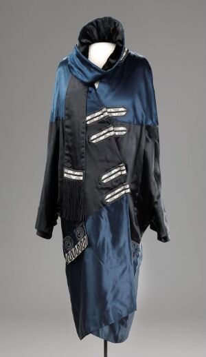  A mannequin wearing an ornate, knee-length midnight blue coat with metallic silver piping and embellishments, featuring an oversized collar that doubles as a hood, set against a neutral grey background.