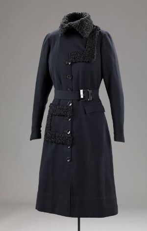  A classic black long-sleeved women's coat with a fur-like collar trim and central button closures, complete with a waist belt and a decorative pocket or flap, presented on a stand against a neutral grey background.