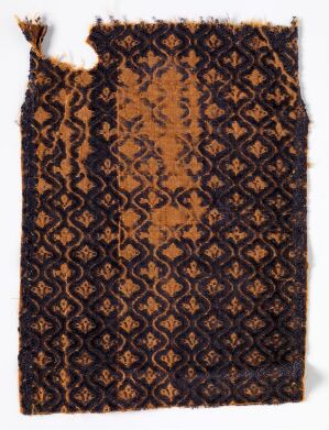  An image of an aged textile fragment with a intricate dark brown or black geometric pattern on a lighter rust-colored background. The textile is frayed on the right edge and torn at the top left corner.