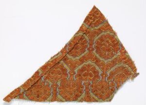  A triangular fragment of fabric with an ornate orange and pale cream floral and vine-like pattern, frayed at the edges, suggesting historical significance and wear.