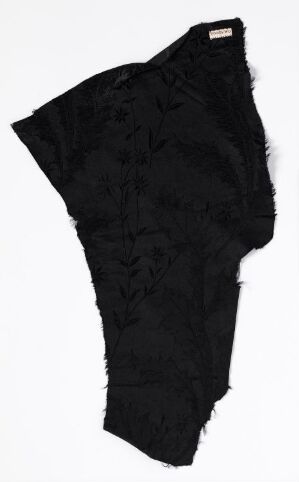  A black, asymmetrical avant-garde garment with ruffled embellishments on one side, displayed against a stark white background. The unique fashion piece has one sleeve and a diagonally cut torso, showcasing an unconventional design.