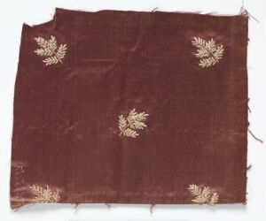  A fragment of aged, maroon fabric with rough edges and sporadic pale botanical motifs, showing signs of wear and with a textured look suggesting historical value. Artistname and title are unknown.