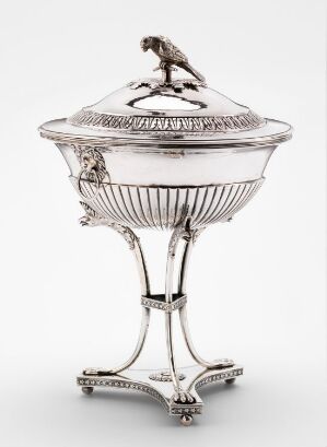  An ornate silver pedestal bowl with a fluted design and a bird figurine on its lid, set against a plain gray background.