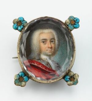  A miniature portrait titled "Johan Georg Büchler" by an unidentified artist, featuring an 18th-century male in formal attire within a round gilded silver frame, detailed with enameled blue flowers with yellow centers and green leaves. The portrait is protected by a clear, edge-cut glass.