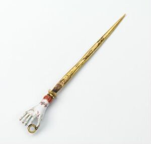  A decorative object with a white, hand-shaped base with red and gold trim, extending into a long, golden, ornately patterned tapering rod or scepter, set against a light background.