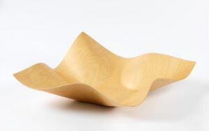  "Tuisku" by Petri Vainio, a laminated and bent plywood tableware piece with a flowing, wavelike design in a light honey brown color set against a plain white background showcasing the natural wood grain.