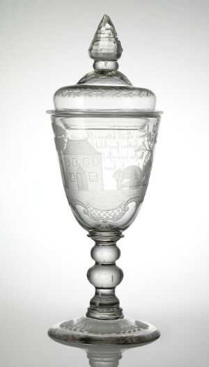  A clear glass goblet with an intricately etched design and a pointed lid, exuding elegance and featuring reflective and frosted patterns. The goblet has a floral-patterned base, a stem with sphere-like elements, and a detailed bowl and lid, showcased against a neutral background.