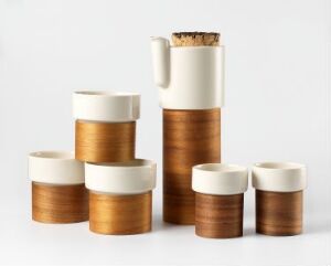  "Warm" tableware collection by Tonfisk Design Oy, with a tall porcelain and walnut veneer pitcher with a cork stopper surrounded by various sized cylindrical containers, showcasing a contrast of matte white porcelain and warm walnut wood textures against a white background.