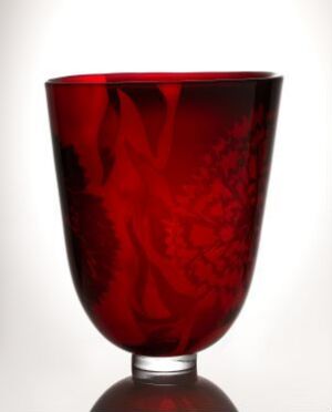 
 A deep red blown glass vase with etched decor titled "Carnation Icelands" by artist Kerttu Nurminen, exhibiting a rich crimson color that fades towards the base and featuring intricate organic patterns etched into the glass.