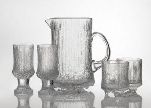  A set of glass tableware designed by Tapio Wirkkala, including one textured pitcher and three textured glasses with a fine vertical pattern, elegantly displayed against a white-to-light-gray gradient background with reflections beneath each piece.