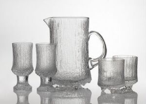  A set of glass tableware designed by Tapio Wirkkala, featuring a textured pitcher and three matching glasses on a reflective surface against a white background, capturing light and showcasing detailed craftsmanship.
